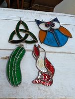 Four stained glass decorative pieces arranged on a white wooden surface including a green triquetra, owl, pickle, and bird shapes.
