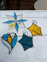 Top down view of four stained glass hanging ornaments on a white surface: an eight-point star in blue and clear, two diamond shapes with wire swirl designs in yellow and teal, and one blue and yellow diamond shaped piece.