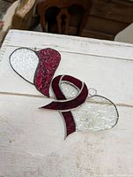 Two stained glass decorative pieces laid on a white wooden surface: one heart shaped and one looped ribbon shape, both featuring deep red and white textured glass.