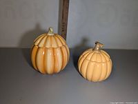 Two ceramic pumpkin-shaped candle containers, one larger with a rounded stem lid, and one smaller, lighter orange with a curved stem lid.
