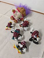 Photo showing the troll doll with purple hair and six Raisins figurines in various poses and accessories on white surface.