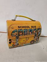 Front and side view of vintage metal school bus lunch box with Disney character illustrations and yellow handle