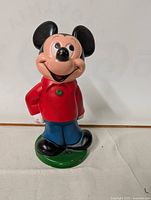 Front view of vintage Mickey Mouse acrylic piggy bank standing on a green base.