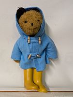 Full view of Paddington Bear doll standing, showing blue coat with hood and yellow rubber boots.