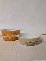 Two vintage Pyrex glass baking dishes, one golden yellow with white floral pattern and one cream-colored with black floral pattern, side by side.