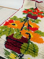 Close-up image showing plush hooked yarn texture and colorful detail of potted flowers in orange and red with green leaves.