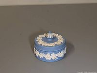 Blue round Wedgwood jasperware container with white floral relief on the sides and lid, lid closed showing finial top