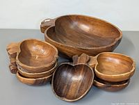 View of the entire set showing one large apple-shaped wooden bowl and six smaller matching apple-shaped wooden bowls, all with carved leaf and stem detailing.