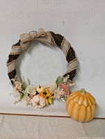 Front view of the fall wreath showing artificial flowers, burlap ribbon, and faux pumpkins on twig base, alongside the ceramic pumpkin candle with lid on.