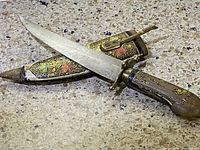 Image showing the dagger out of its holder, displaying the blade, painted wooden handle with gold inlay, and brass guard.