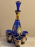 Photo of a cobalt blue glass decanter with stopper and six matching shot glasses, featuring gold hand-painted floral motifs on all pieces.