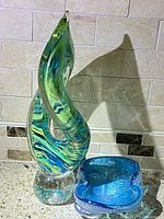Photo of the two glass sculptures on a kitchen counter with tan tile backsplash showing the tall twisted green and blue sculpture and the smaller blue dish or ashtray.