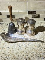 Four vintage items on a terrazzo countertop: a metal bell with wooden handle, two silver-finished shakers likely salt and pepper, and an intricately engraved decorative spoon.