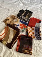 Seven folded scarves displayed together showing various colors and mixed patterns lying on a white quilted surface.