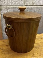 Closed teak wood ice bucket with lid on, showing wood grain exterior and one ring handle with metal accent.