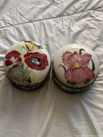 Two closed oval porcelain trinket boxes with floral and butterfly designs on lids, placed on a white fabric surface