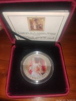 Colorized Canadian 25 cents coin featuring Queen Elizabeth II in a protective capsule inside a red-lined black presentation box with paperwork behind it.