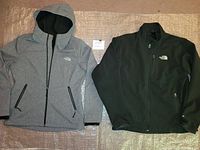 Front view of both jackets, grey women's hoodie on the left and black men's APEX jacket on the right, showing logos and front details