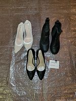 Set of three women's shoes displayed on a bubble wrap surface: white fishnet pumps, black wedge booties, and black pumps.