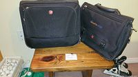 Two black Swissgear bags on a wooden table; one expandable messenger bag with 'benefits today' patch, one wheeled computer business case.