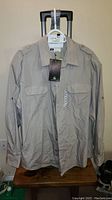 Front view of men's khaki outdoor shirt showing long sleeves, buttoned chest pockets, shoulder epaulets, lightweight fabric with tag attached.