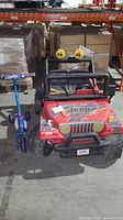 Photo showing red Jeep Wrangler style kids ride-on car and purple/blue Huffy scooter side by side indoors in warehouse setting.