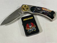 Photo showing open folding pocket knife with stainless steel blade and handle featuring a motorcycle motif and gold emblem, alongside a black lighter with detailed 'American Biker' graphic.
