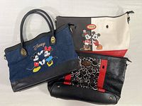 Three Disney tote bags displayed together: one denim with handles featuring Mickey and Minnie embroidery; one red, black, and white tote with printed Disney characters, no handles; and one black with red stripes tote decorated with Mickey patterns, no handles.