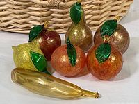 Photo showing six of the seven hand blown art glass fruits in front of a woven basket: pear, apple, lemon, peach, orange, tangerine, and banana on table.
