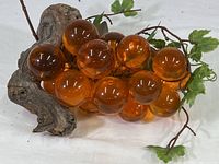 Front angle showing the cluster of translucent orange and amber acrylic grapes attached to a twisted natural wood grape root stem with green plastic leaves and vine branches.