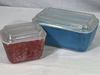 Two rectangular Pyrex covered dishes side by side, one red and one blue, both with clear glass lids, showing overall appearance and color condition.
