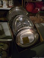 Six silverplate trays stacked in a box showing varied sizes and decorative details.