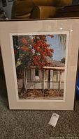Framed art print featuring an old house with a bright red-orange flowering tree in front, sitting on carpeted floor.