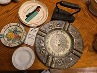 Top-down view showing antique iron, large metal clock face ashtray, and four smaller ashtrays on wooden table surface.
