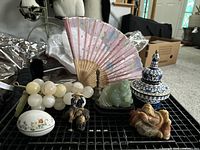 Overview of lot including two carved Asian figurines, carved stone grapes, cloisonne lidded dish, ceramic figurine, folding hand fan and metal bell on black tray