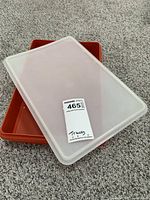 Rectangular red Tupperware container with semi-transparent white lid on carpet.