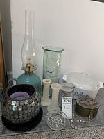 Eleven assorted vintage houseware items displayed, including oil lamp, candleholders, brass box, purple candle on black plate, clear jars, apothecary jar, ashtray, and beaded bookmark.