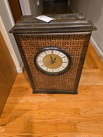 Wooden clock with wicker front design on hardwood floor, showing entire clock front and structure.