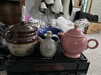 Brown and cream ceramic bean pot, pink Fiesta Ware teapot, small gray pottery oil pitcher, blue mixing bowl, and beige mixing bowl arranged on floor