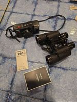 Olympus camera, Microwave Ranger binoculars, and Bleu de Chanel cologne sample on blue surface