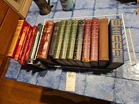 Photo showing 14 vintage books including red hardcover bibles, green leather-bound book volumes, and other hardcovers on a shelf rack.