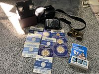 Overview of lot showing Canon camera with strap on carpet beside black Hama camera case, antique brass binoculars, Sawyers View-Master viewer, and 10 View-Master reels arranged in two rows, along with Westinghouse Magic Flash Cubes box.
