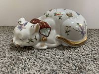 Side view of white ceramic sleeping cat figurine decorated with colorful butterflies, flowers, and foliage, showing the red collar and gold details.