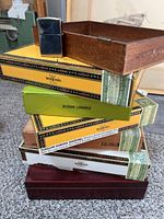 Stack of six cardboard cigar boxes mainly Don Tomas brand, a wooden humidor box on top and a silver Zippo lighter placed on wooden humidor