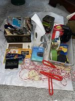 Overview of the lot showing white portable clothes steamer, Amazon Echo Dot box, multiple sewing-related items in a tin, household aids like Command strips, twine, brushes and other miscellaneous items.