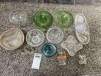 Assortment of vintage glassware including green glass juicers, cut glass bowls, a marbled bowl, clear and colored glass containers, and glass salt and pepper shakers arranged on a carpet.