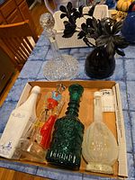 Full view of lot showing clear cut-glass stoppered decanter, various decorative glass bottles including dark green embossed bottle, white opaque bottle, and dark glass vase with artificial black flowers arranged on a blue tiled table.