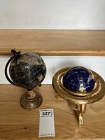 Two globes on wood surface: black globe with bronze-style stand and blue lapis ocean gemstone globe on tripod stand.