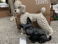 Full view of white ceramic poodle statue standing behind a smaller black terrier statue on carpeted floor with cardboard box background.