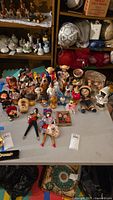 Wide view of assortment of dolls on table with shelves behind, showing different styles and cultural costumes.